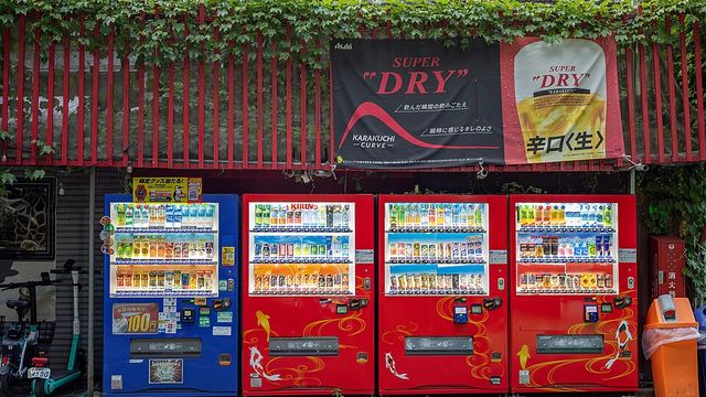 A row of four drinks vending machines under a shop awning, with a recycling bin for cans at the right-hand end.