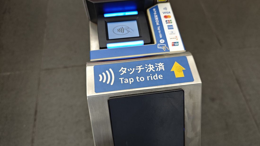 A ticket gate’s contactless reader with a blue “タッチ決済 / Tap to ride” sticker below it and a yellow arrow pointing up at the panel.