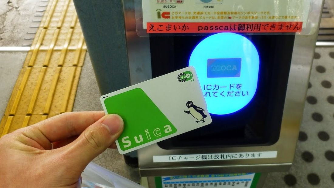 A hand holding a Suica card flat against the glowing blue reader of a platform validator.